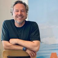 Markus Wenzel – Gastgeber mit Leidenschaft bei A&M Sylt Mann sitzt vor A&M-Sylt-Plakat mit Urlaubsbotschaft.