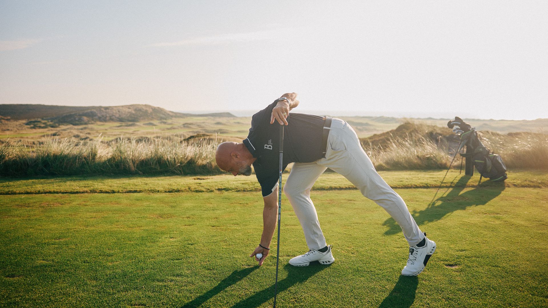  Mann legt Golfball auf den Rasen, Golftasche im Hintergrund.