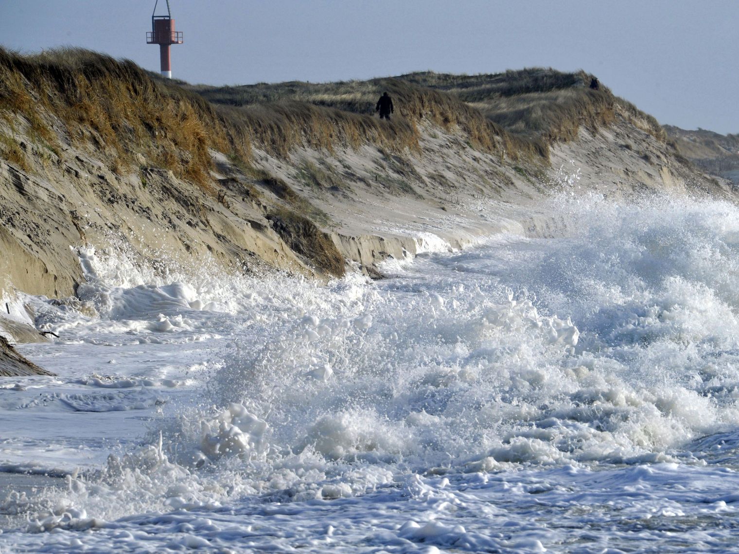Hohe Wellen schlagen schäumend gegen eine steile Dünenküste.