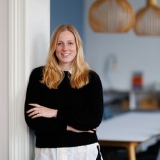 Eine Frau mit langen blonden Haaren steht lächelnd an einer weißen Wand in einem modernen Büro. Sie trägt einen schwarzen Pullover und hat die Arme vor der Brust verschränkt. Im Hintergrund sind zwei große hölzerne Designer-Lampen und ein Besprechungstisch mit Stühlen zu sehen.