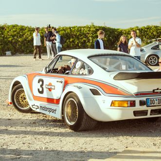 Weißer Porsche im Martini-Design auf sandigem Parkplatz mit Menschen.