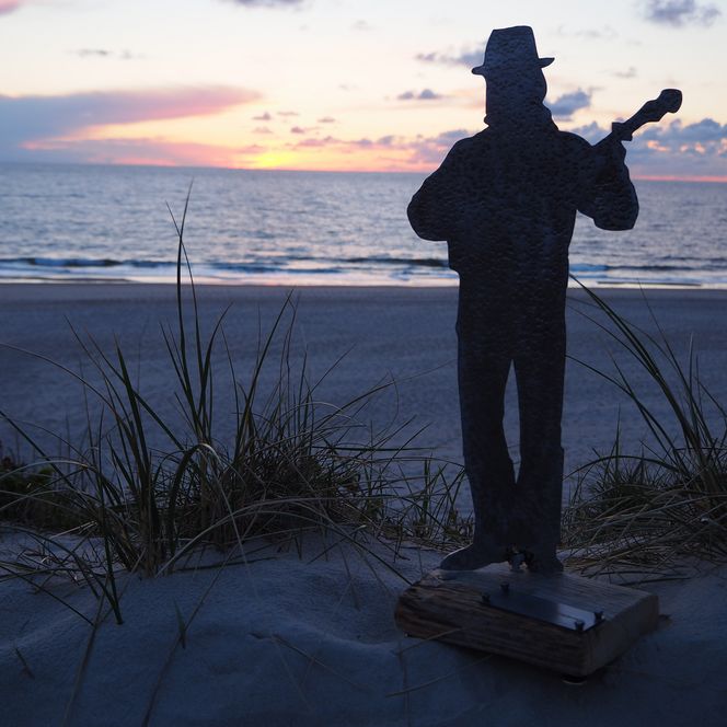 Skulptur „Der Henner“ vor Dünen, Strand und Sonnenuntergang.