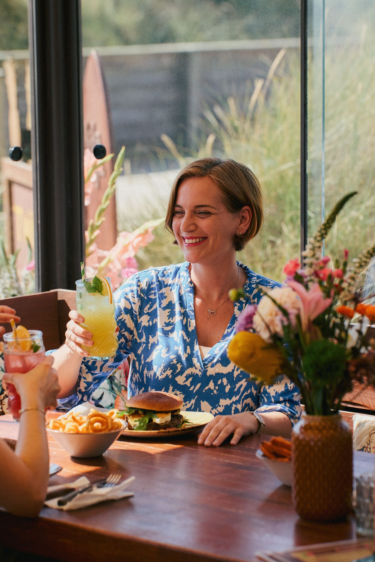 Eine Frau in einem blau-weißen Kleid sitzt im Twisters Beach Dinner in Wenningstedt auf Sylt, hält ein Cocktailglas und lächelt an einem Tisch mit Burger, Pommes und Blumen.