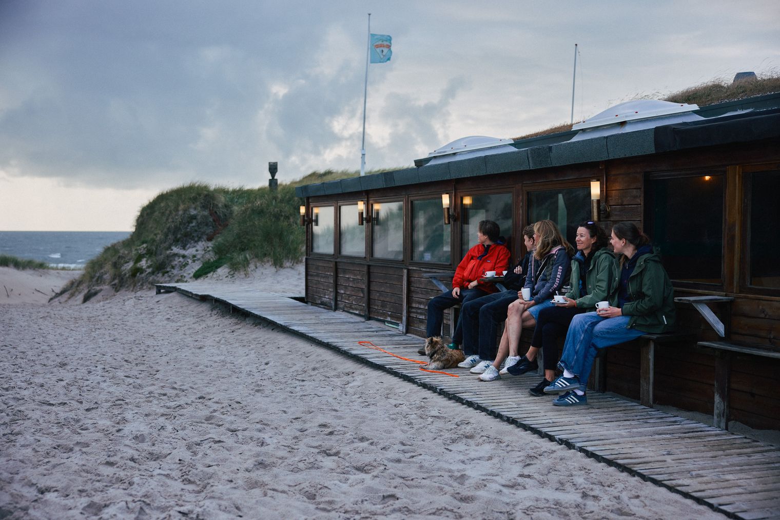 Fünf Personen sitzen mit Getränken vor einer Strandhütte auf einer Holzbank, neben ihnen liegt ein kleiner Hund. Im Hintergrund sind Dünen und das Meer unter einem bewölkten Himmel.