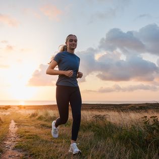 Eine Frau joggt auf einem Pfad durch die Natur, während die Sonne am Horizont aufgeht.