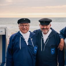 Drei Männer in maritimen Uniformen und Mützen lächeln vor einem Strandkorb, mit Meer und Sonnenuntergang im Hintergrund. 