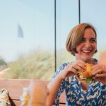Donna che ride mentre mangia un hamburger in un ristorante con vista sul prato e sul cielo azzurro.