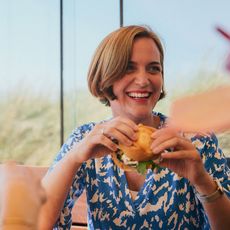 Lachende Frau isst einen Burger in einem Restaurant mit Ausblick auf Gräser und blauen Himmel.