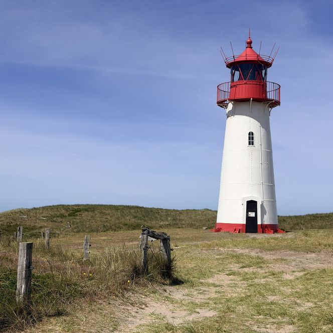 Weißer Leuchtturm mit rotem Dach auf einer grünen Düne. Ein Sandweg und ein Holzzaun führen durch das Gras. Der Himmel ist blau mit leichten Wolken.