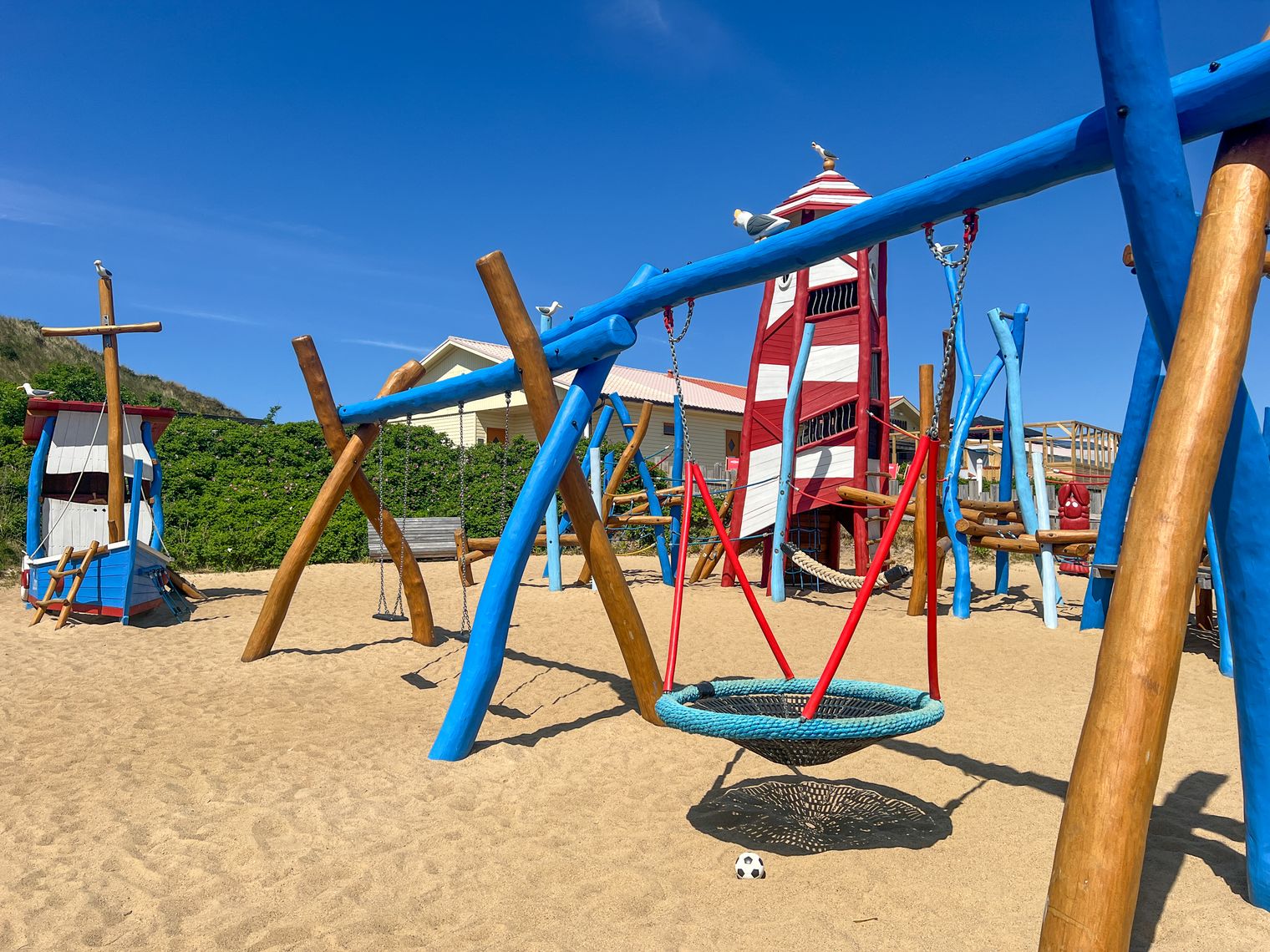 Bunter Spielplatz mit Leuchtturm, Holzschiff und Schaukeln auf Sandboden.