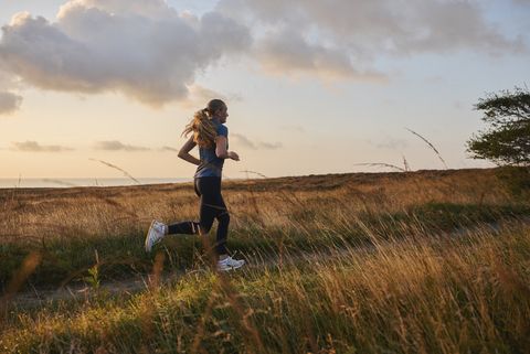 Eine Frau läuft bei Sonnenaufgang durch eine Wiesenlandschaft, während Gräser im Vordergrund wehen.