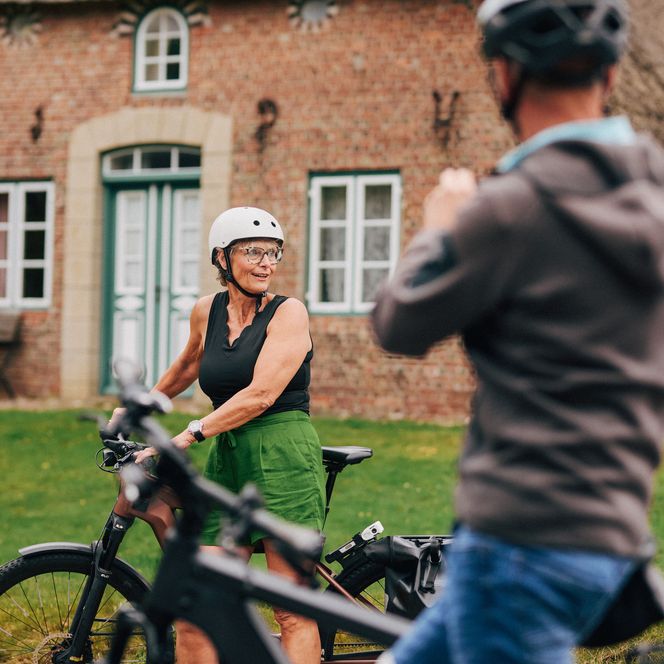 Ein Mann und eine Frau auf E-Bikes in Keitum auf Sylt vor einem historischen Reetdachhaus, beide lachen und tragen Helme.