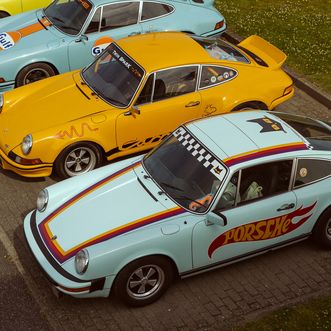 Mehrere farbige Porsche-Oldtimer stehen nebeneinander auf einem Parkplatz.