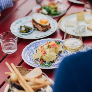 Verschiedene Speisen, darunter Käse, Kartoffeln und Brot, auf einem rot lackierten Tisch im Käseklub in Keitum auf Sylt, mit Gläsern Wein und Wasser.