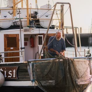 Fischer auf Boot repariert Netz, Hafen und Wasser im Hintergrund.
