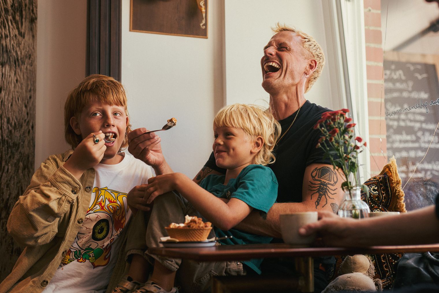 Ein Vater mit zwei Kindern sitzt in einem gemütlichen Café. Ein Kind isst Kuchen, das andere sitzt auf dem Schoß des Vaters, der herzlich lacht. Auf dem Tisch stehen Kaffee und eine Vase mit Blumen.