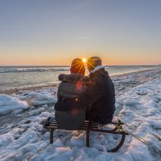 Ein Paar sitzt auf einem Schlitten am verschneiten Strand von Hörnum auf Sylt und genießt den Sonnenuntergang über dem Meer.