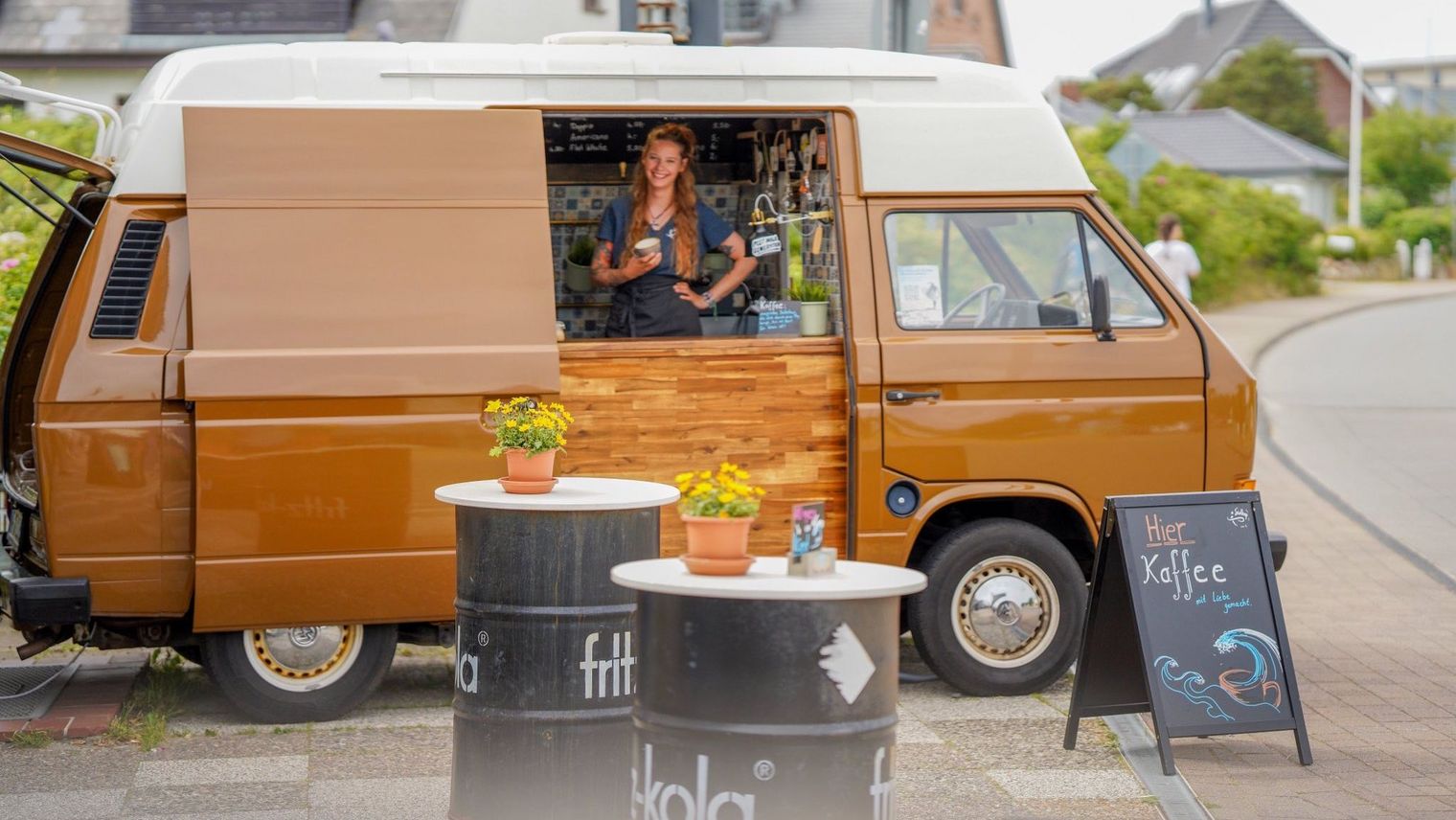 Frau serviert Kaffee aus einem umgebauten VW-Bus mit Stehtischen davor.