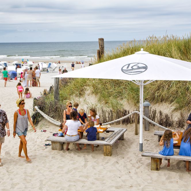 Menschen sitzen unter einem weißen Sonnenschirm an Holztischen im Sand, im Hintergrund Strandkörbe und Meer.