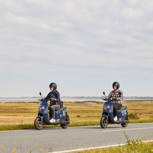 Zwei Menschen fahren auf blauen E-Rollern an der Sylter Küste entlang, mit Wattenmeer im Hintergrund.