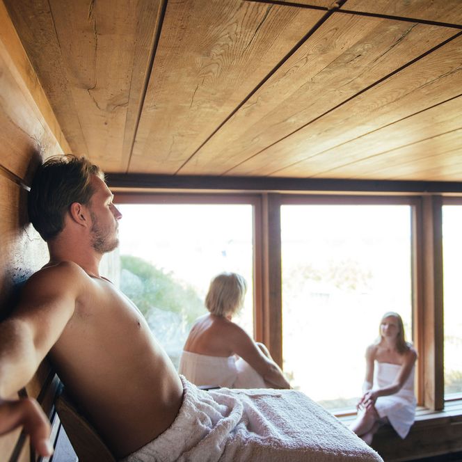 Sauna in der Sylter Welle Ein Mann sitzt entspannt in einer Blockhaussauna mit Blick durch Panoramafenster, während eine Frau und ein Mädchen im Hintergrund die Aussicht genießen.