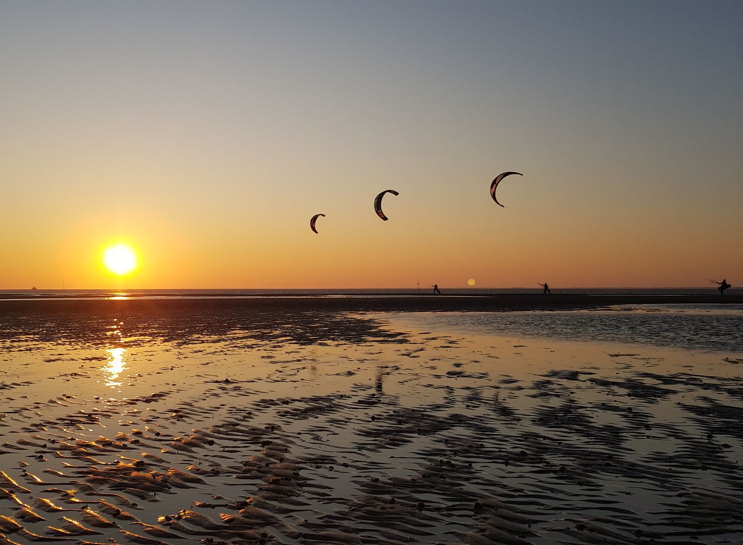Drei Kitesurfer gleiten bei Sonnenaufgang über das flache Wasser vor Hörnum auf Sylt.