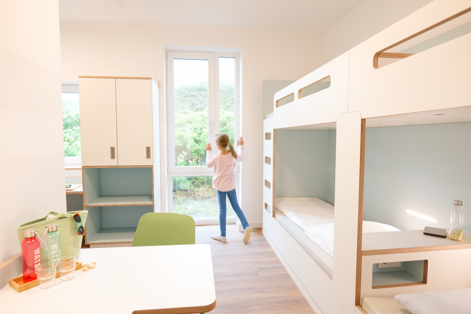 Kind schaut aus Fenster in hellem Vierbettzimmer mit Etagenbetten und Schränken.