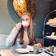 Eine junge Frau genießt eine Tasse Tee in einem gemütlichen Cafe, vor sich eine Etagere mit Gebäck und Sandwiches.