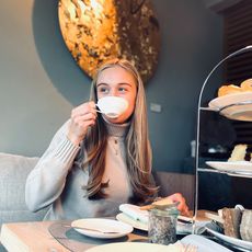 Teezeremonie im Kontorhaus Keitum Eine junge Frau genießt eine Tasse Tee in einem gemütlichen Cafe, vor sich eine Etagere mit Gebäck und Sandwiches.
