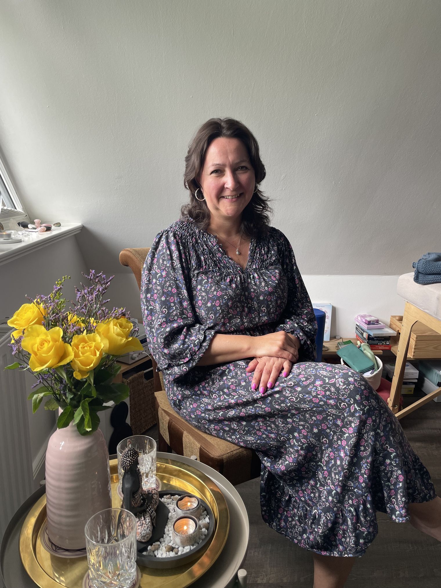 Eine Frau sitzt lächelnd im geblümten Kleid neben gelben Rosen und Kerzen.