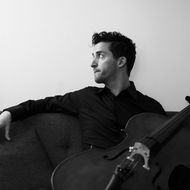 Mann sitzt mit Cello auf Sofa und blickt nach rechts.