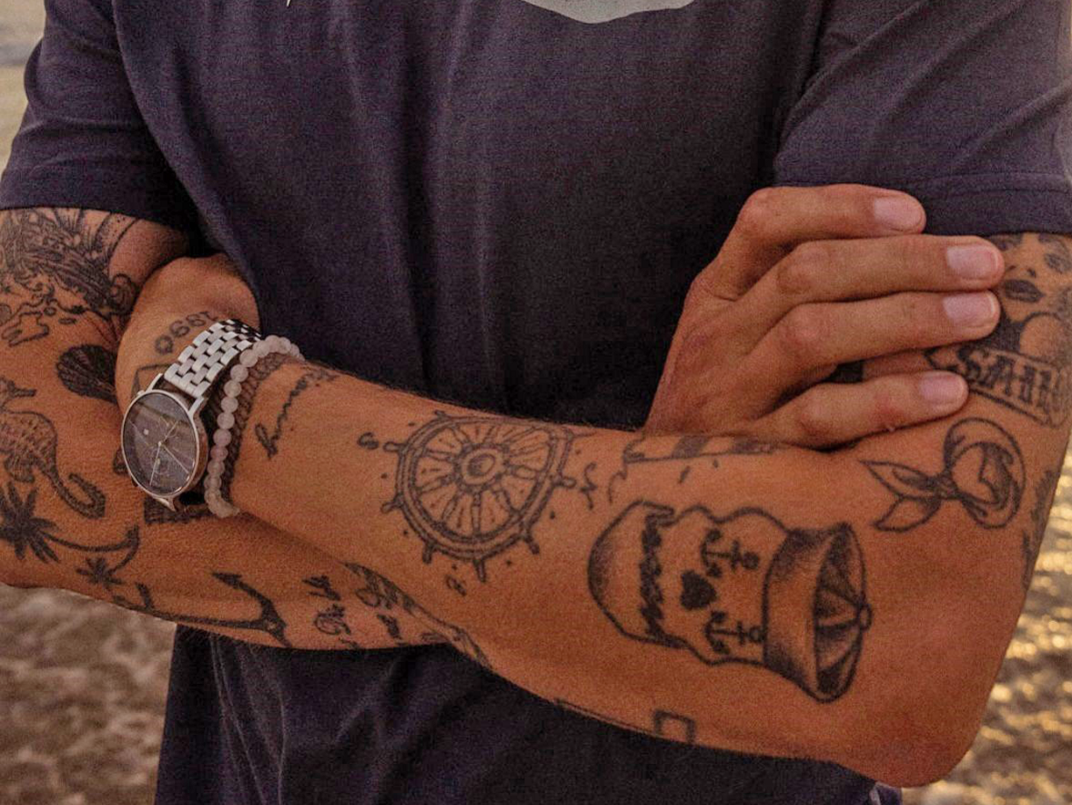 Verschränkte, tätowierte Arme vor Meer, dunkles T-Shirt mit Samoa Logo.