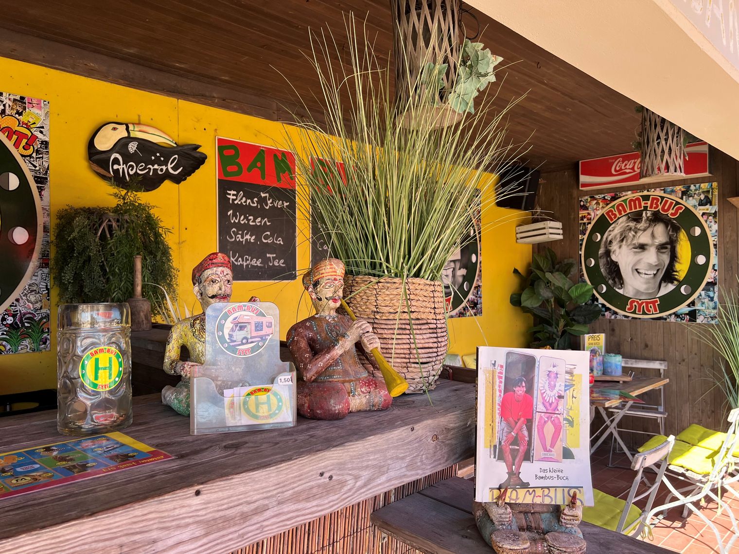 Alternativer Text Bunte Bar-Dekoration mit Figuren, Pflanzen, Stickern und Retro-Postern.