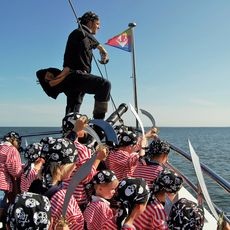 Eine Gruppe von Kindern in rot-weiß gestreiften Piratenkostümen mit schwarzen Kopftüchern steht auf dem Deck eines Schiffes. Sie halten Spielzeug-Säbel hoch. Ein Erwachsener, als Pirat verkleidet, steht auf der Reling mit einem Säbel in der Hand. Die Nordsee erstreckt sich im Hintergrund unter einem klaren blauen Himmel.