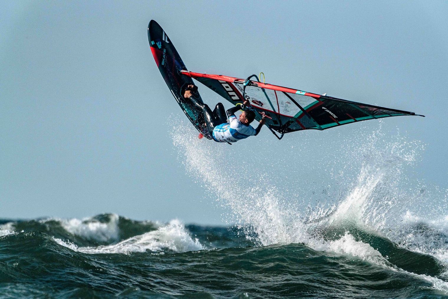 Windsurfer springt mit Board über Welle bei Wind und Gischt.