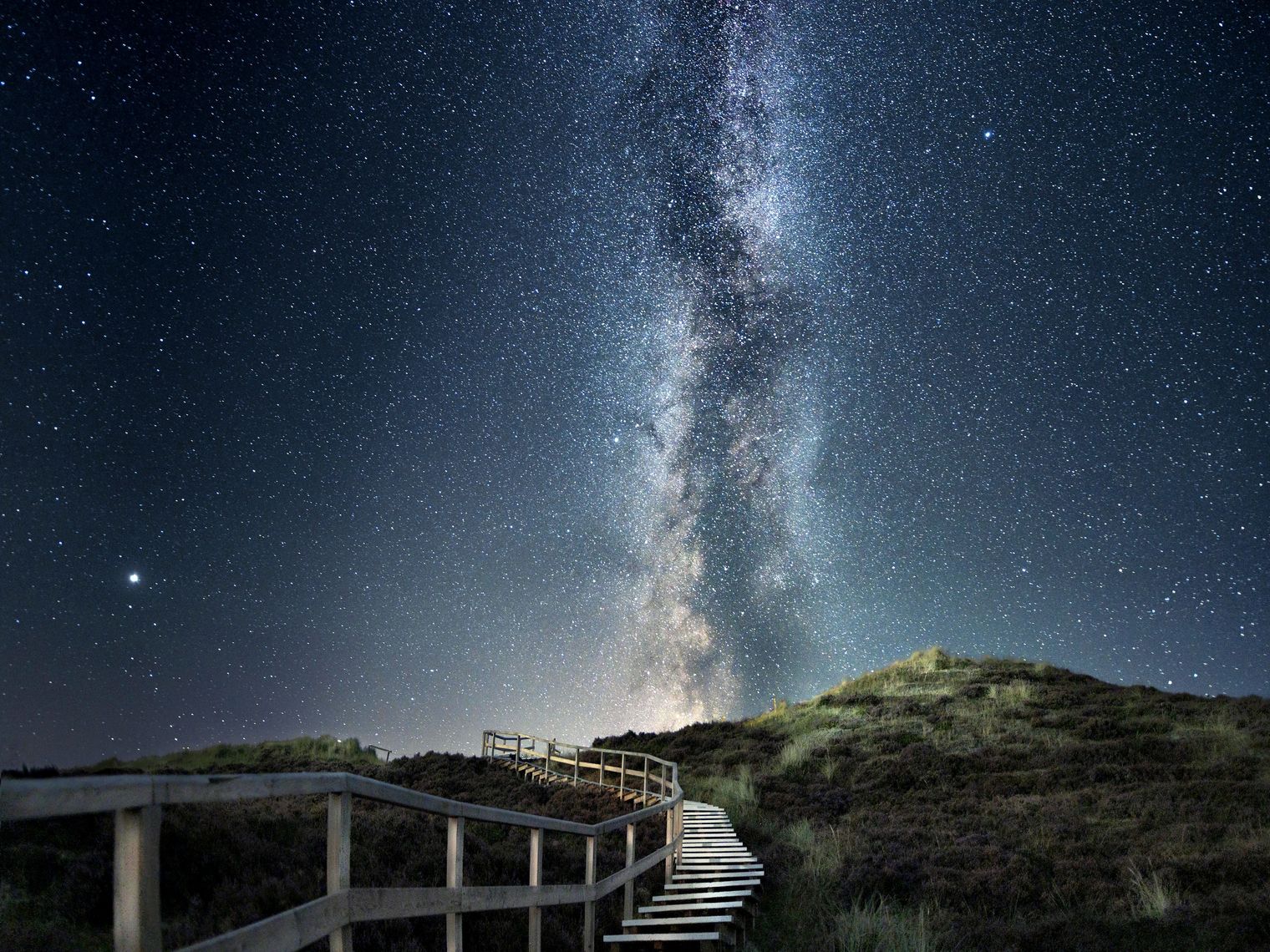 Holzsteg durch Dünen unter einem klaren Sternenhimmel mit Milchstraße.