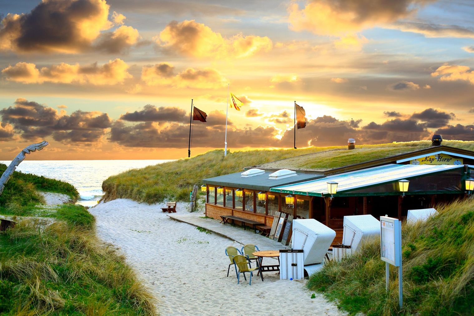 Strandrestaurant Kap-Horn zwischen Dünen mit Sonnenuntergang im Hintergrund, Strandkörbe und Flaggen davor.