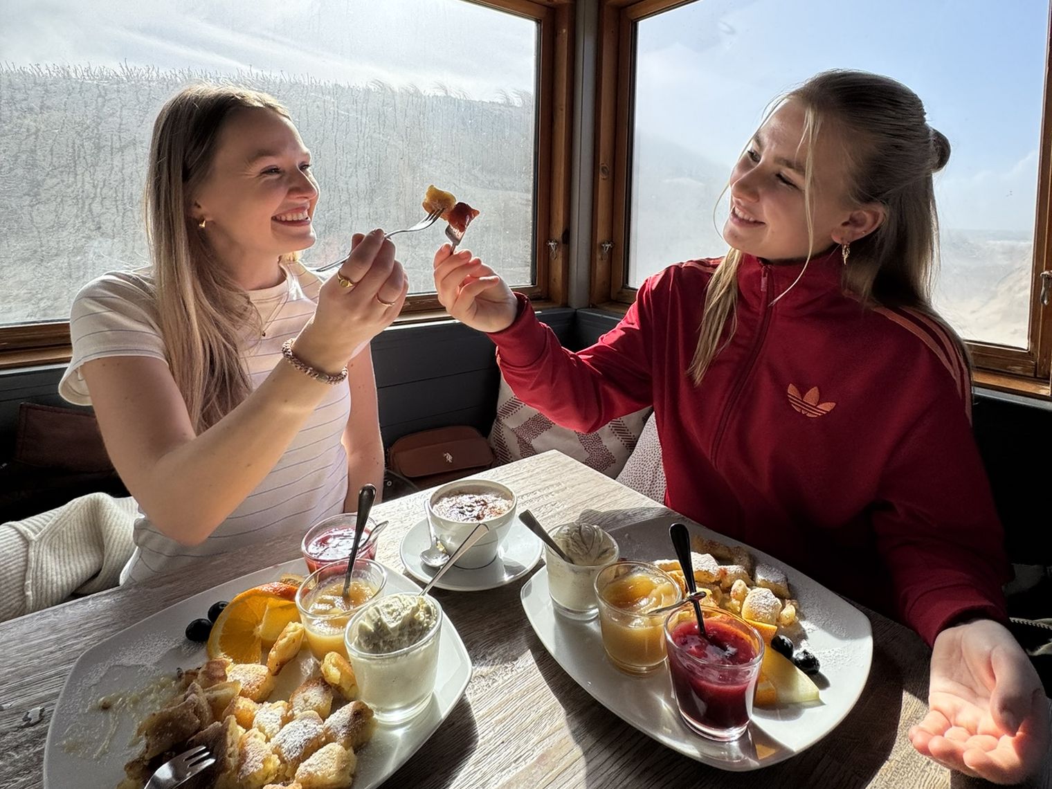 Zwei Frauen teilen sich an einem Holztisch Kaiserschmarrn in einem rustikalen Restaurant mit großen Fenstern.