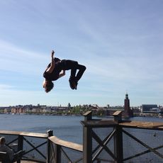 Eine Person macht einen Rückwärtssprung in der Luft über einer Uferpromenade mit Stadt und Wasser im Hintergrund.