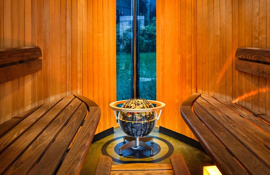 Fährhaus Spa Sauna mit Holzbänken, einem modernen kugelförmigen Saunaofen und großem Fenster mit Ausblick auf eine grüne Landschaft.