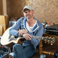 Mann mit Baseballcap und Gitarre sitzt auf Stuhl und blickt in die Kamera.