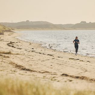 Eine Frau läuft am Sandstrand entlang, die Weite des Meeres und die sanften Hügel der Dünenlandschaft bilden eine ruhige Kulisse.