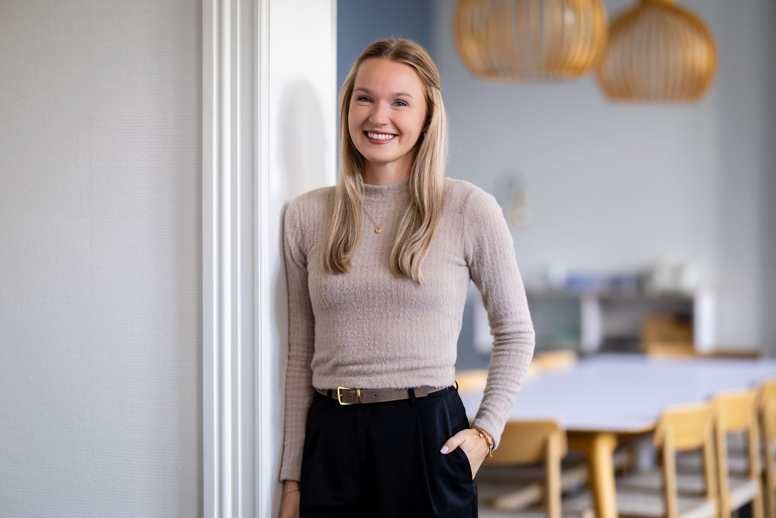 Eine junge Frau mit blondem, offenem Haar trägt einen beigen Pullover und eine schwarze Hose, steht lächelnd an einer weißen Wand vor einem modern eingerichteten Raum.