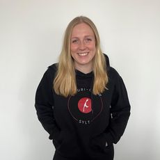 Eine junge Frau mit blonden Haaren steht vor einer neutralen weißen Wand und lächelt. Sie trägt einen schwarzen Kapuzenpullover mit dem Logo der Azubi-Crew Sylt 2024 und hat ihre Hände in den Taschen.