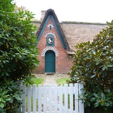 Charmantes Reetdachhaus mit grüner Tür, Rhododendren und weißem Picketzaun.