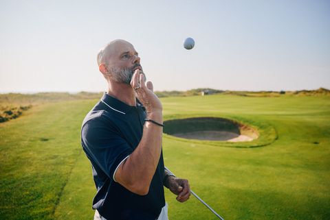 Golfer fixiert fliegenden Ball mit Golfbunker im Hintergrund.