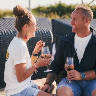 Lachendes Paar mit Aperitifgläsern auf Sonnenterrasse.