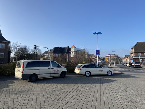 Zwei Taxis stehen an einem Taxistand vor dem Bahnhof in Westerland. Im Hintergrund sind Gebäude und eine Bushaltestelle zu sehen, der Himmel ist blau und klar.