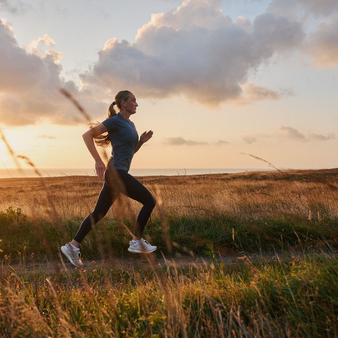 Joggerin in schwarzer Sportkleidung läuft bei Sonnenuntergang durch eine grasbewachsene Dünenlandschaft an der Nordsee.