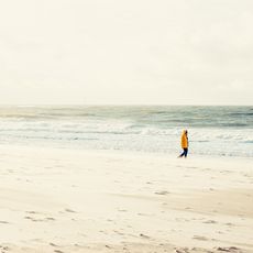Eine Person in einem gelben Regenmantel spaziert am breiten Sandstrand entlang, während im Hintergrund Wellen unter einem wolkigen Himmel zu sehen sind.